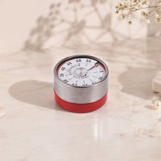 Round Cre8tion mechanical timer with a steel dial face and a red base, standing on marble.